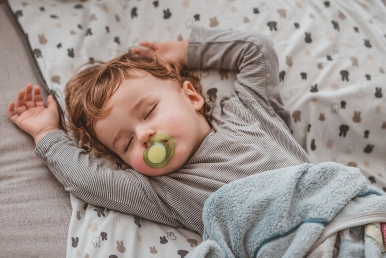 A young child with light brown hair sleeps serenely on a bed adorned with white sheets featuring small animal designs. Wearing a gray long-sleeve shirt, the child has a green pacifier in their mouth and is covered with a blue blanket, arms stretched above their head—a picture-perfect scene for any sleep consultant.