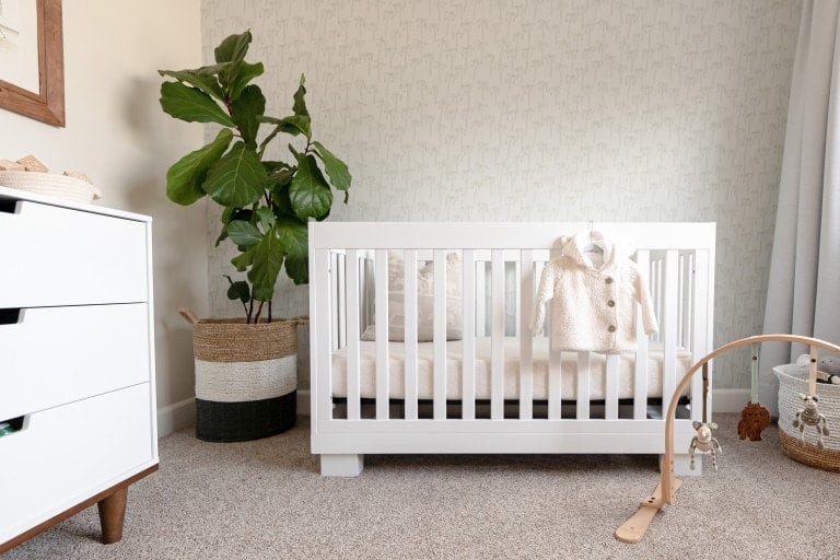 A minimalist, eco-friendly nursery featuring a white crib with a knit outfit hanging on it. Nearby is a large houseplant in a woven basket, a white dresser with drawers, and a wooden baby gym with toys on the carpeted floor. The wall has subtle patterned wallpaper.