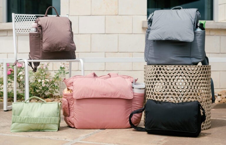 The image shows five Calpak baby bags of various sizes and colors displayed outside. A brown backpack is placed on a white chair, while a green bag, pink bag, and black bag are on the ground. A gray backpack-like bag is placed on a perforated stool.