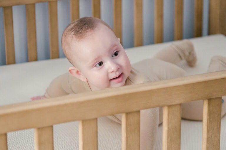 A baby in a beige outfit is lying on their stomach in a wooden crib with light-colored bedding, resting comfortably on a Naturepedic mattress. The baby is looking to the side, appearing curious or engaged, with the crib bars visible in the foreground.