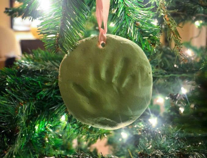 A handmade Christmas ornament featuring a green clay handprint hangs from a branch of a Christmas tree, illuminated by small white lights. The ornament is suspended by a pink ribbon, making it an ideal Christmas ornament for toddlers.