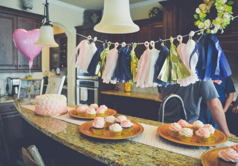 A kitchen island decorated for a birthday theme celebration with pink and gold cupcakes on plates, a large frosted cake, and a garland with blue, white, and gold tassels hanging above. Two pendant lights are overhead, casting a warm glow on the heart-shaped pink balloon. Twinkle twinkle little star accents complete the look.