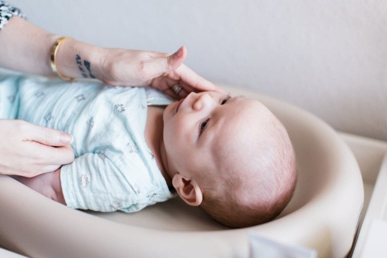 A baby wearing a light blue onesie lies on a soft, cushioned surface. An adult hand gently touches the baby's mouth, possibly offering relief from baby constipation. The baby has short, light-colored hair and pale skin. The adult is wearing a gold bracelet and has a tattoo on their forearm.