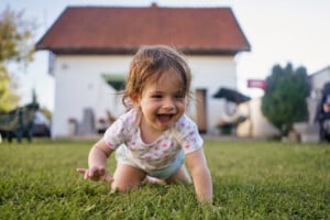 A toddler with brown hair is crawling on a grassy lawn in front of a white house with a red roof. The young girl, perhaps named Lily or Leah, is wearing a white shirt with colorful patterns and has a joyful expression on her face. Trees and outdoor furniture are in the background.