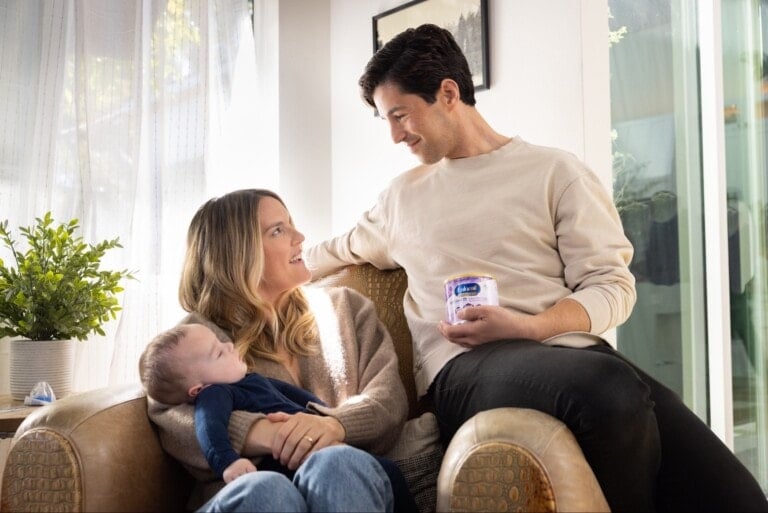 A person is seated on an armchair holding a baby. Another person, who resembles Josh Peck, stands beside them, holding a container and smiling at the seated person. There is a potted plant and window with curtain in the background. The scene appears indoors.
