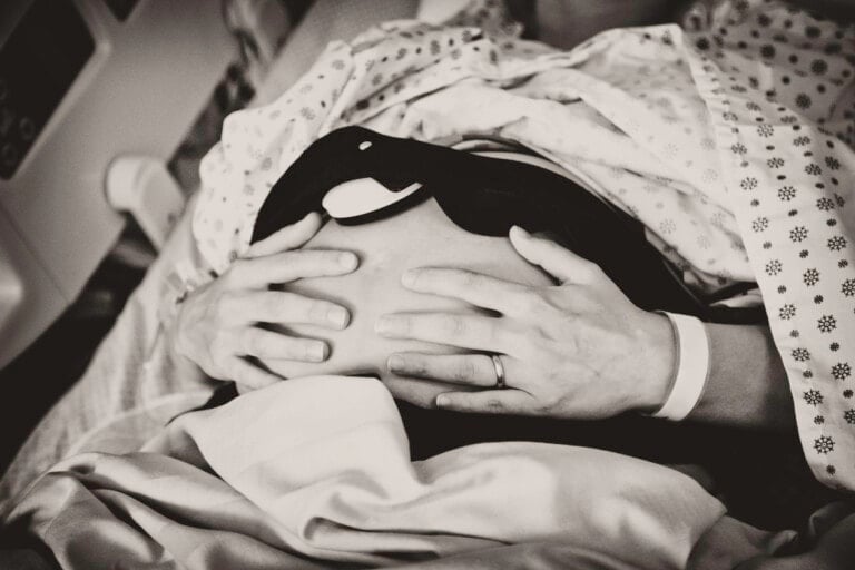 A person is lying on a hospital bed, wearing a patterned gown. They are resting hands on their exposed, pregnant belly, displaying a hospital wristband and a ring. The image in black and white poignantly captures the silent fear of potential complications like placental abruption.