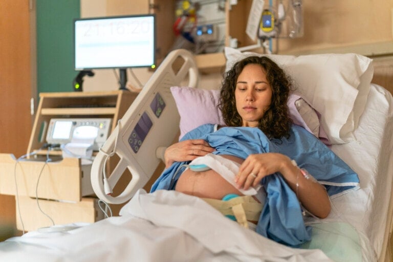 A pregnant woman lies in a hospital bed, wearing a blue hospital gown. Medical monitors and equipment are visible beside her as she rests her hands on her belly, with a fetal monitor attached. The room, which appears to be a maternity ward, is prepared for potential labor induction methods.