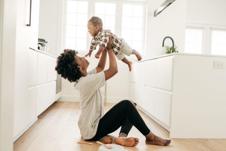A minimalist mom is sitting on a kitchen floor, lifting a baby above her head. The well-lit kitchen features white cabinets and large windows. She’s wearing a grey shirt and black leggings, while the baby sports a plaid shirt and diaper.