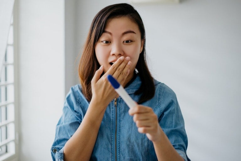 A woman with shoulder-length hair wearing a denim shirt looks at a pregnancy test result with an expression of surprise, covering her mouth with one hand while holding the test with the other. She wonders, "When is the Best Time to Take a Pregnancy Test?" as she stands indoors near a window.
