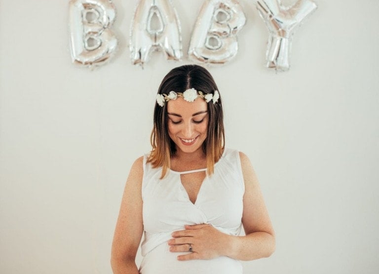 A pregnant woman in a white dress stands with her hands resting on her belly, smiling. She is wearing a flower crown and standing in front of silver balloon letters that spell "BABY." The scene radiates joy, like the excitement felt during the best baby shower games.
