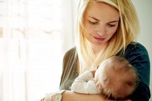 A woman with long blonde hair is holding an infant in her arms near a sunlit window. She looks down at the baby with a gentle expression. Wearing a watch and a dark top, she epitomizes self-care for moms, ensuring both of them are enveloped in love as the baby is wrapped in a light-colored blanket.