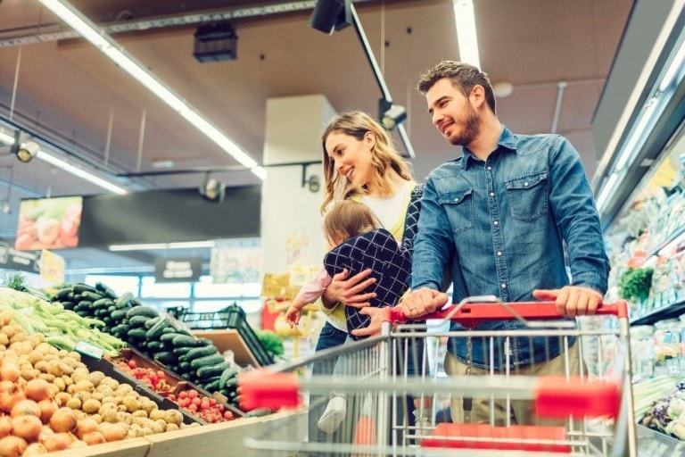 A couple with a small child shop for produce in a grocery store, mindful of their grocery budget. The woman holds the child while the man pushes a shopping cart. They are looking at vegetables on display, including zucchinis and potatoes. The store has bright lighting.