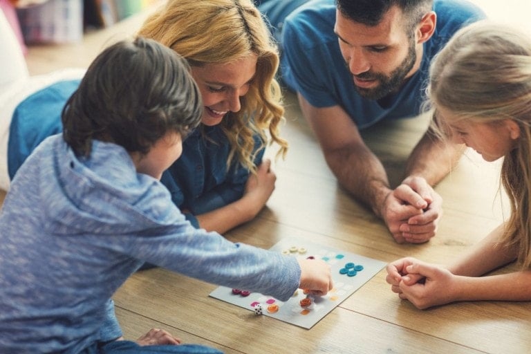 Two adults and two children are lying on the floor and playing a family game together. The children, a boy and a girl, appear engaged in the game while the adults, a man and a woman, look on and participate. They are in a brightly lit room with wooden flooring.