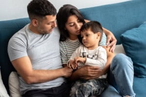 A man, woman, and child sit closely huddled on a blue couch, empathy evident in their warm embrace. The man wears a gray t-shirt, the woman a striped shirt, while the child sits on her lap in a white shirt with dark pants.