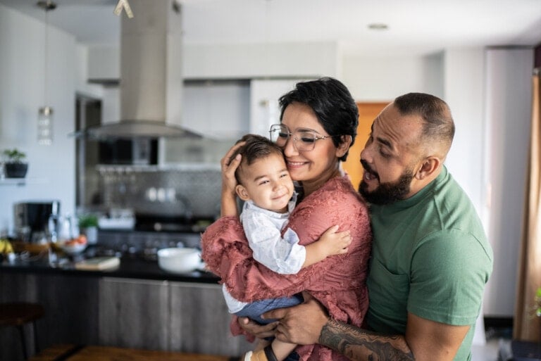 A woman wearing glasses holds her smiling only child, a toddler, as a man stands close by, also smiling at the child. They are in a modern kitchen with various appliances and items on the counters in the background.