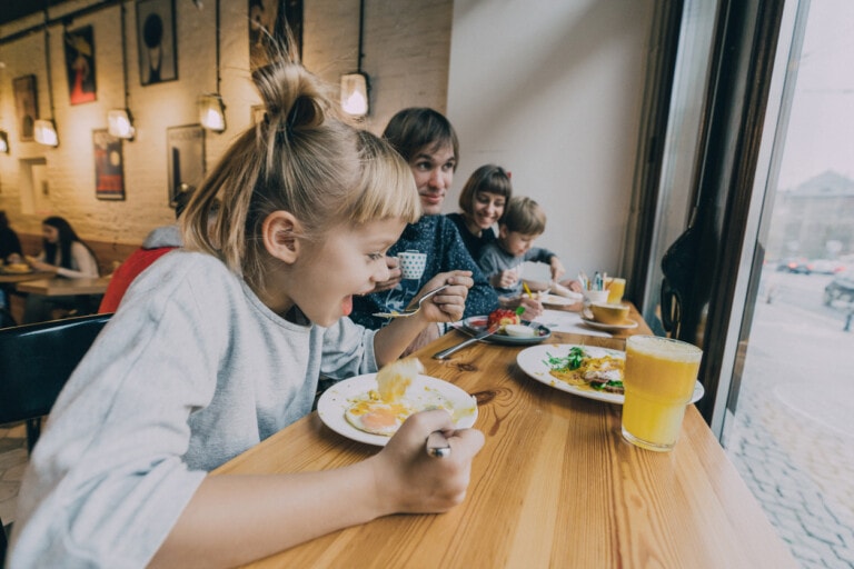 A family of four is shown eating at a wooden table by the window in a family-friendly restaurant. Two children are in the foreground, one eating, while a mother and another child are seated behind them. Plates with food and glasses of orange juice are on the table.