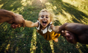 A young child, smiling widely, is being swung around by two adults holding her hands. She is wearing a yellow dress and appears to be enjoying the playful activity in an outdoor grassy area—one of many wonderful things to do while your child is little.