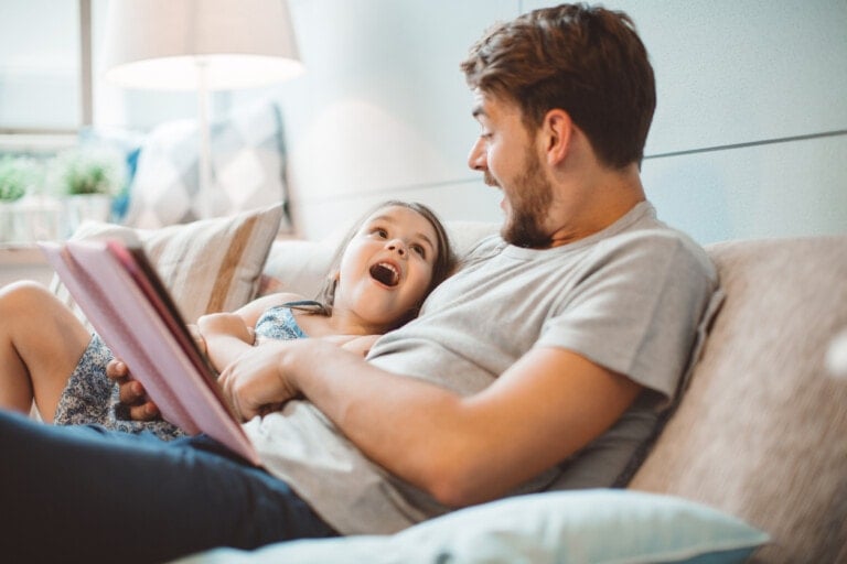 A man and a young girl sit on a couch. The man, perhaps one of those dads acting as babysitters, holds a book or tablet and looks at the girl who has an excited expression on her face. The scene unfolds in a cozy living room with a lamp and pillows in the background.