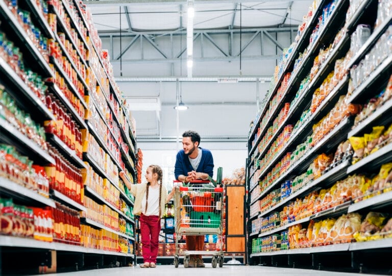 A man and a child are shopping in a supermarket, pushing a shopping cart with the bare minimum essentials down an aisle lined with various colorful products on shelves. The child is pointing at something on a shelf while the man looks on, smiling. The store has bright lighting and a high ceiling.