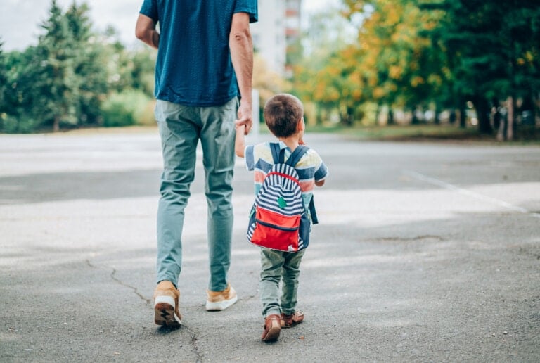 A man and a young child holding hands and walking outdoors on a paved surface. The child, who is sporting a colorful striped backpack filled with back to school must-haves, walks beside the man dressed in casual clothing. The area has trees and greenery in the background.