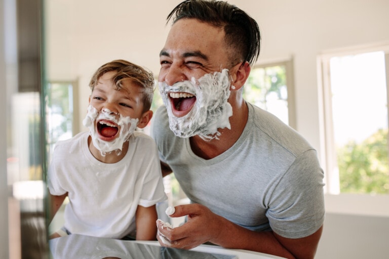 A father and son with shaving cream on their faces are laughing together in a bathroom. The man is wearing a gray t-shirt, and the boy is in a white t-shirt. They appear to be enjoying a playful moment.