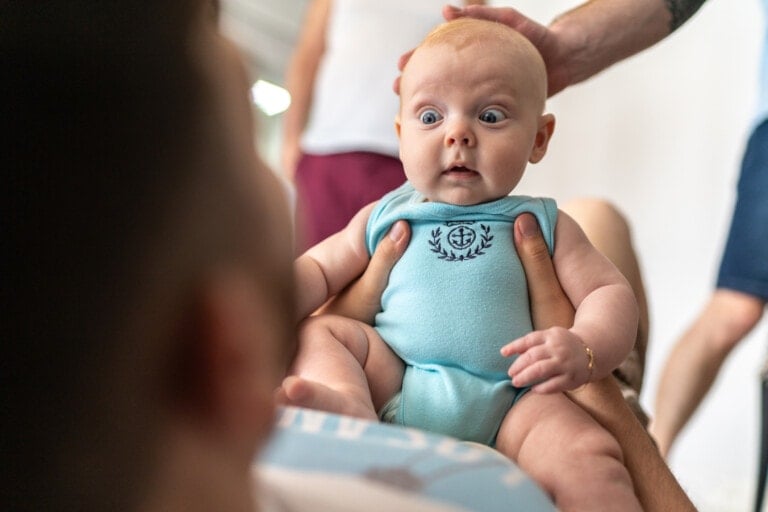 A baby wearing a light blue onesie with an anchor symbol is held by an adult. The baby has wide eyes and an open mouth, appearing surprised or curious—a moment that captures the essence of parenting mistakes. There are blurred figures and objects in the background.