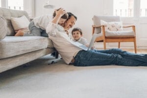A man lies on the floor using a laptop, while a child hugs him from behind on the sofa, and another child sits beside him smiling. The room's light-colored interior features a chair and a cushion in the background, creating a cozy atmosphere that encourages everyone to behave better.