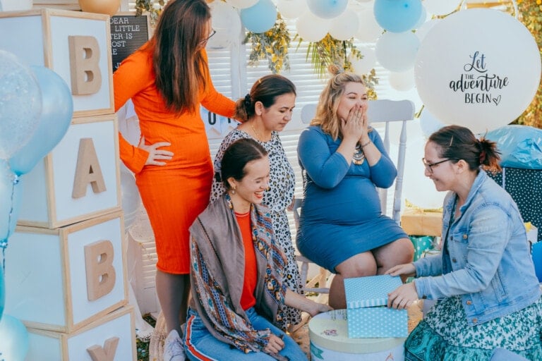 Five women are gathered at a baby shower with delightful baby shower themes. One woman, wearing a blue dress, is visibly pregnant and covering her mouth in delight. Another woman is holding a gift box. The area is decorated with balloons, a 'BABY' sign, and a 'Let the adventure begin' sign.
