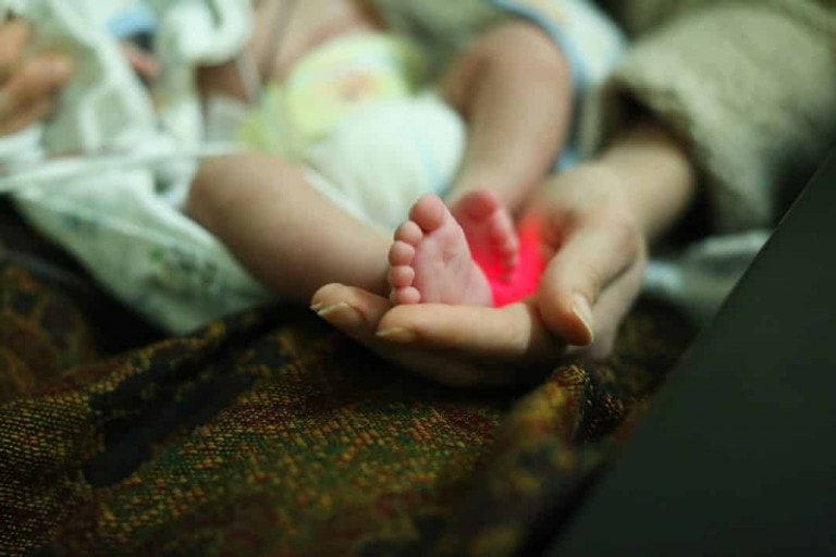 A close-up image of an adult hand gently holding a small NICU baby's foot. The red light around the baby's foot suggests medical monitoring or a health check. The background is blurred and includes a blanket or fabric.