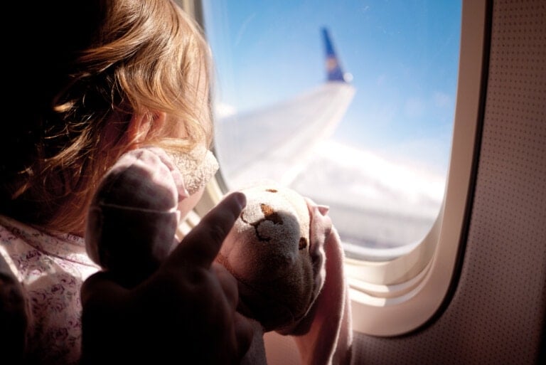 A child holding a stuffed animal is looking out the window of an airplane. The light shines on the child and toy, with a clear blue sky visible beyond the plane's wing. Nearby, a small bag of airplane snacks for kids adds to the scene’s charm.