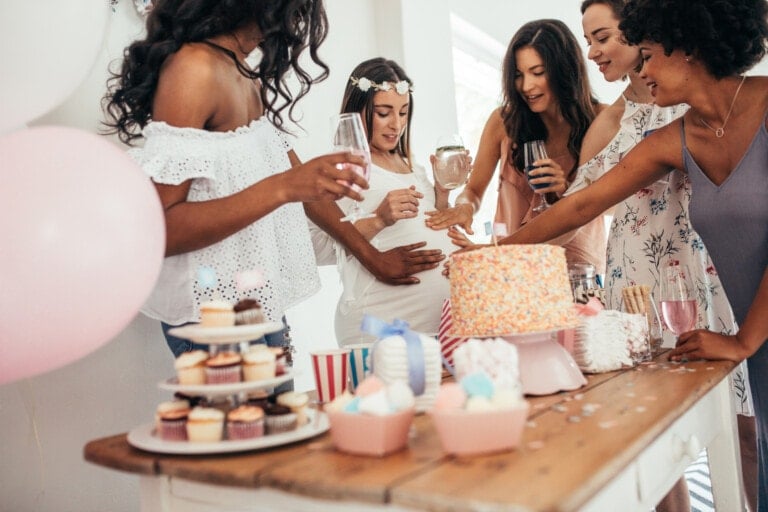 A group of six women stand around a table filled with baby shower food ideas, including cupcakes and a large cake. One woman in the center, who appears to be pregnant, is being celebrated. The women are smiling and holding drinks, surrounded by pastel-colored decorations.