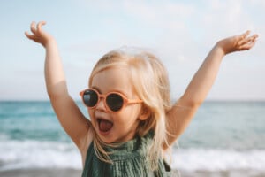 A young child with blonde hair wearing pink sunglasses and a green dress stands on a beach with their arms raised and a wide smile. The ocean is visible in the background with waves gently crashing, capturing the carefree spirit often associated with beautiful girl names.
