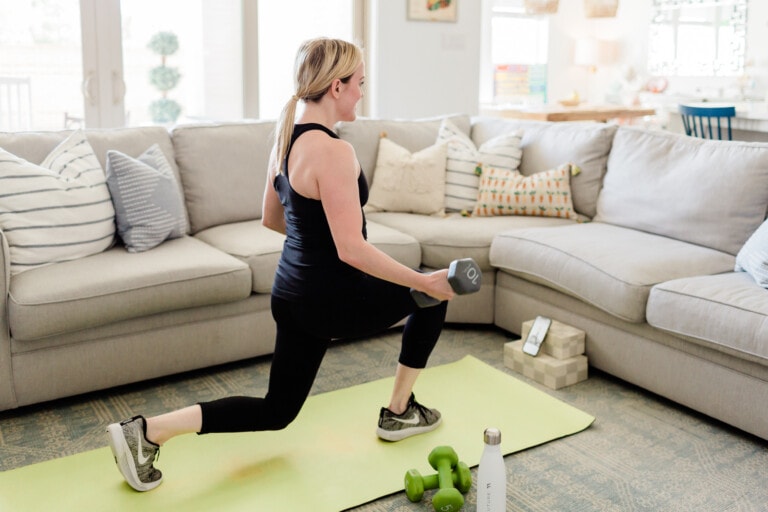 A person with blonde hair in a ponytail is exercising indoors, performing a lunge while holding a dumbbell in one hand. They are on a yellow exercise mat next to a gray couch with decorative pillows. Green dumbbells and a water bottle, essential gear for their FUTURE app workout routine, are nearby.