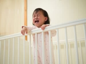 A baby in a pastel pink onesie stands crying in a white crib, gripping the top rail with both hands. The room has soft lighting and a neutral wall in the background. The babys expression shows distress.