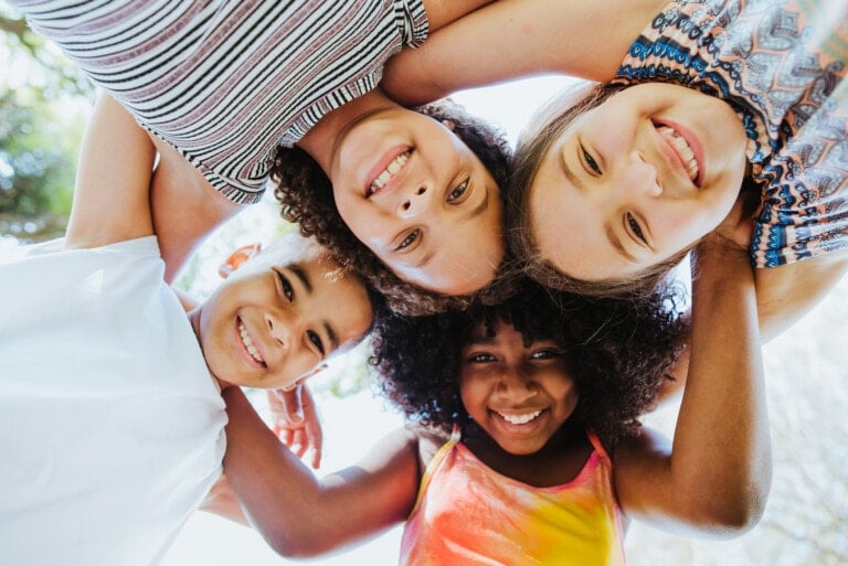 Four children are standing in a circle with their arms around each other's shoulders, looking down and smiling at the camera. They are outdoors, and the background is blurred with green foliage and bright light, capturing a moment of joy that reflects the importance of teaching diversity.
