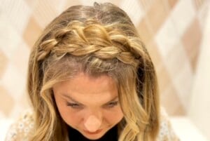 Close-up of a person with blonde hair styled in a braid that wraps around the top of their head, forming a headband-like effect. Ideal among hairstyles for busy moms, the person is looking downward, and the background consists of a light-colored, blurred pattern.