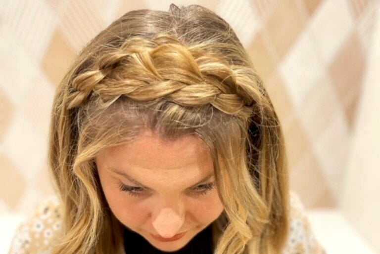 Close-up of a person with blonde hair styled in a braid that wraps around the top of their head, forming a headband-like effect. Ideal among hairstyles for busy moms, the person is looking downward, and the background consists of a light-colored, blurred pattern.