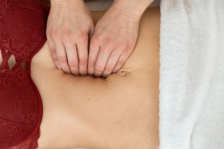 A person is receiving a targeted massage on their abdomen to aid in their diastasis recti recovery. Two hands apply gentle pressure near their navel. The person is wearing a red lace bra and lying on a white towel, focusing on healing abdominal separation.