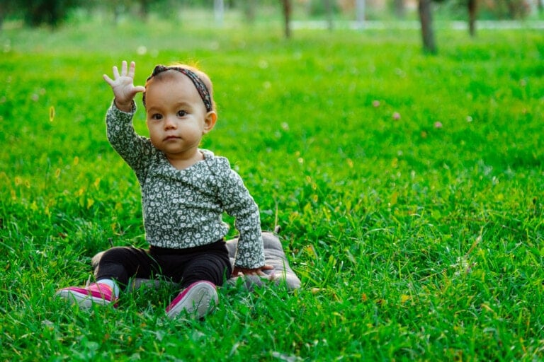 A toddler named Ella sits on the grass in a park, wearing a patterned long-sleeve shirt, black pants, and pink shoes. The child has a headband and is holding one hand up. Trees and greenery are in the background.