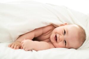 A baby with light skin is lying on a white bed, partially covered by a white blanket. The baby, with one of those rare boy names, is propped up on their stomach, looking at the camera and smiling with an open mouth.