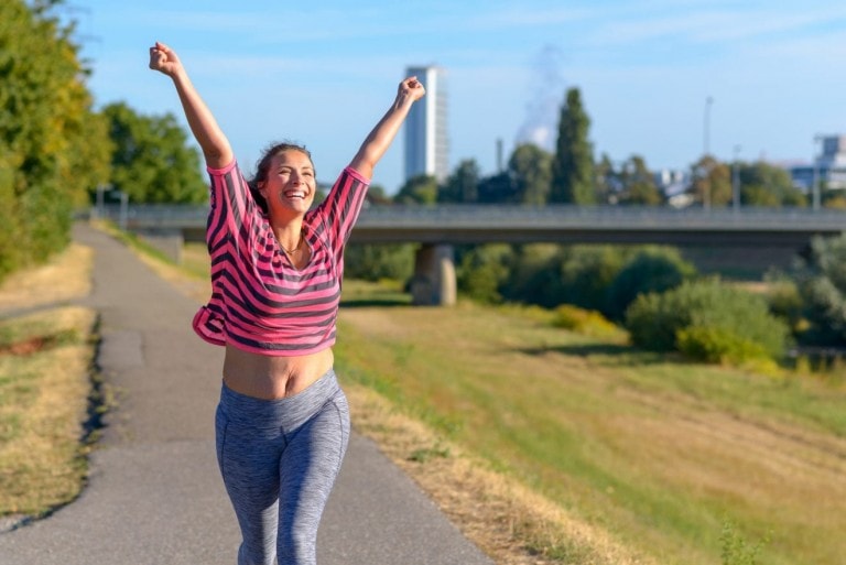 A person dressed in a pink and black striped top and grey leggings is jogging on a paved path with greenery and buildings in the background. They have both arms raised high, smiling broadly under a clear blue sky, radiating the joy of loving their body.