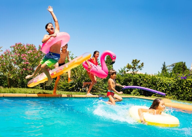 Children are playing in an outdoor pool on a sunny day. One child is jumping into the water holding a float ring, while others are swimming or floating on the best pool toys for kids, including an inflatable pink flamingo. The surroundings include greenery and bright, blue skies.