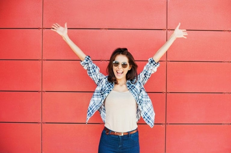 A woman wearing sunglasses, a plaid shirt, and jeans stands in front of a red wall with her arms raised in triumph and a joyful expression on her face, as if she just found everything moms need at Target.