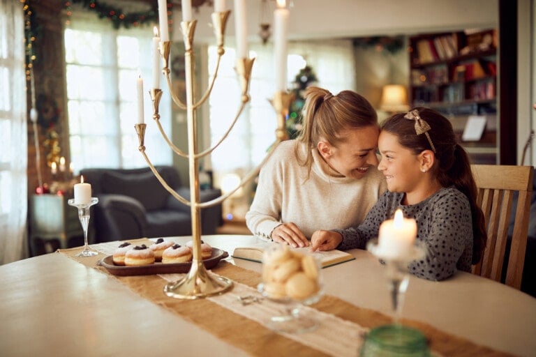 A woman and a young girl smiling and leaning close together while sitting at a table with lit candles, food, and Hanukkah children's books. They are in a cozy, warmly lit room with holiday decorations in the background. The woman appears to be helping the girl with something on the table.