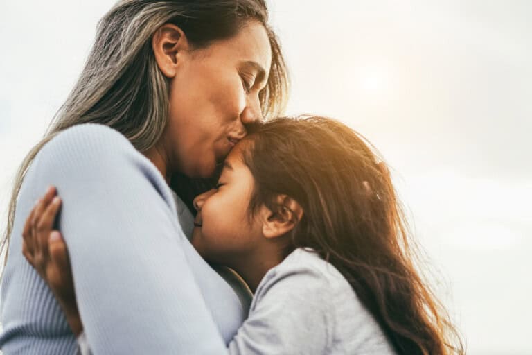 A happy latin mother with long hair embraces her young daughter outdoors, embodying the tender parenting truths. She kisses the top of the girls head, both with eyes closed, while the sun peeks through a bright sky in the background.