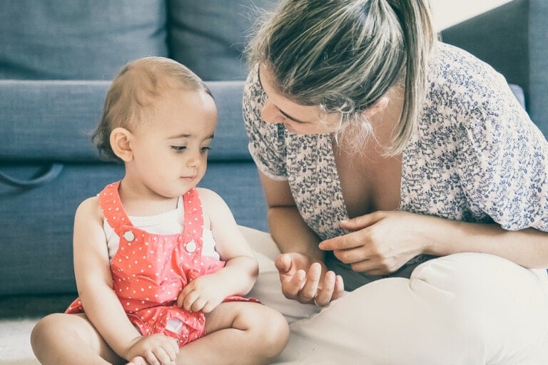 A baby sits on the floor wearing a red dress with white polka dots. An adult with a patterned top and tied-back hair is sitting next to the baby, looking and gesturing towards them, perhaps trying to get kids to listen. They are in a room with a grey couch in the background.