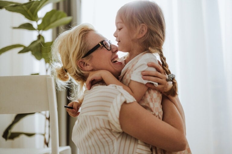 A woman with glasses affectionately holds a young girl with braided hair. Both are smiling as they lean close to each other. The woman wears a striped shirt, and the girl is wearing a patterned dress. In the background, you can see two potted plants and a white chair.