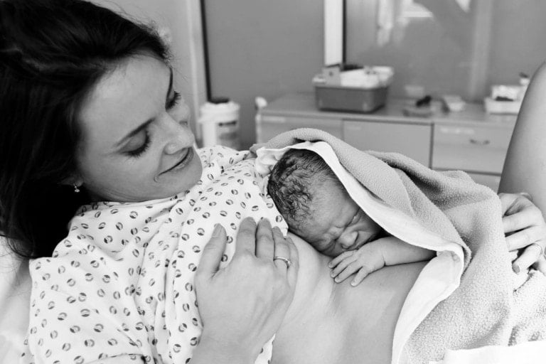 A mother lies in a hospital bed, smiling as she holds her newborn baby on her chest. The baby is wrapped in a towel and appears to be sleeping. The mother, wearing a patterned hospital gown, reflects the joy often discussed on the Healthier Birth Podcast. Medical equipment and supplies are seen in the background.
