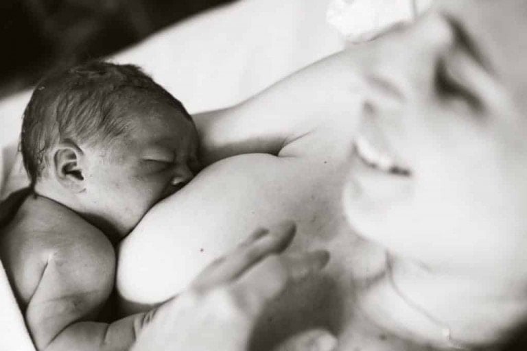 A black and white image showing a newborn baby breastfeeding. The baby has its eyes closed, latched onto the mother's breast to receive colostrum benefits. The mother is smiling and laying on her side, cradling the baby.
