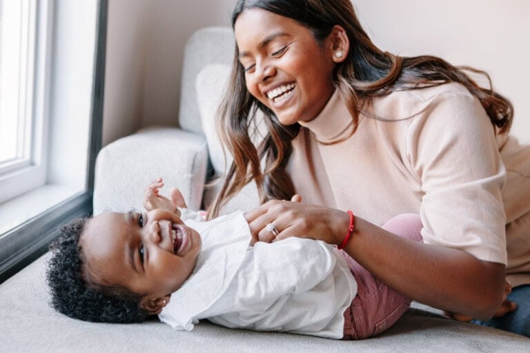 An adult woman leans over and smiles as she tickles a laughing baby who is lying on a couch near a window. The baby is wearing a white shirt and pink pants. Both appear happy and engaged.